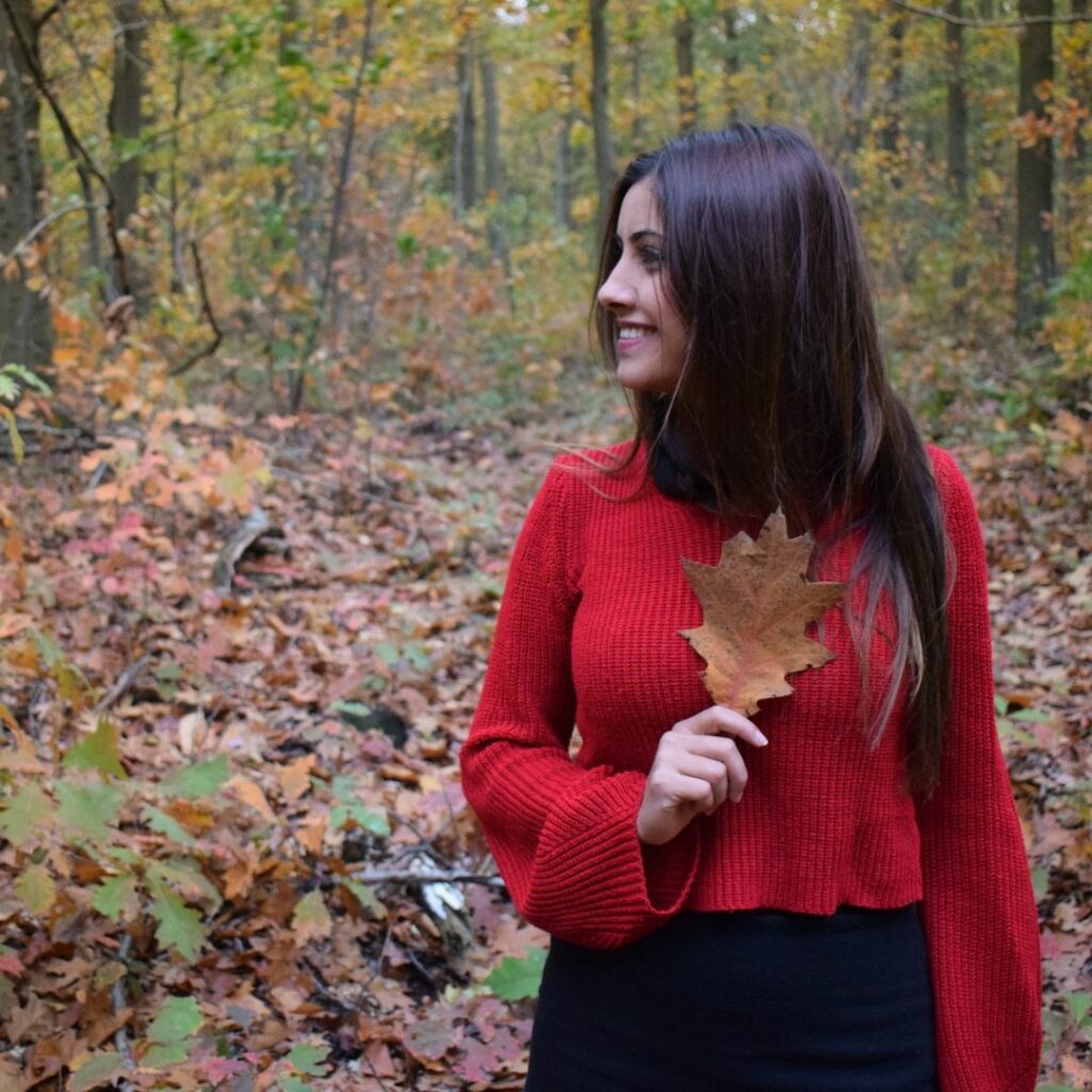 Chica joven con un jersey rojo en medio de un bosque de otoño. Lleva una hoja cogida en la mano y sonríe mirando a un lado. En El Formiguer tenemos primeras marcas de tiendas de moda del Berguedà, ¡y puedes comprarlo online!