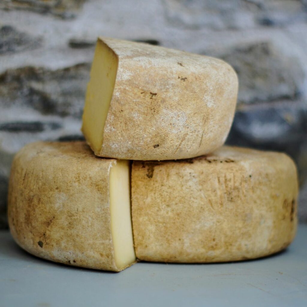 Un formatge artesà, amb la pell tacada pels ferments, amb una cunya del mateix formatge a sobre, davant una paret de pedra. Troba aquí el formatge dels nostres productors!